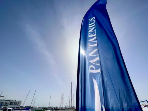 Beachflag mit dem Logo von Pantaenius im Wind, Segelbootmasten im Gegenlicht im Hintergrund